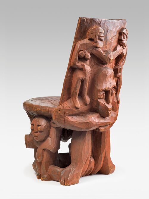 A detail from a photograph of a wooden chair with an intricately carved backrest. Several figurative designs are carved into the wood, including human figures arranged in a group. The sculptures are sculpted in relief from the solid wood.