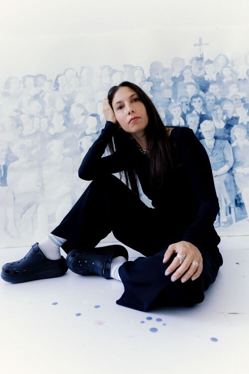 The artist Giulia Andreani is sitting on the floor in front of a large blue illustration featuring shadowy figures. She is wearing dark clothes, white socks and black shoes; one leg is bent and she is resting her head on one hand.