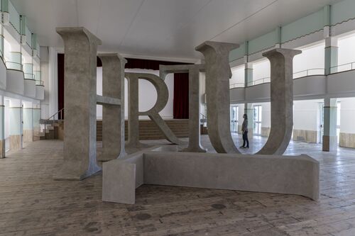 A large-scale sculpture comprising monumental, stone-coloured letters H, R, T and U in a spacious indoor hall. The oversized letters are arranged in such a way that visitors can walk through them. Tall windows can be seen in the background. A single person walks past the letters in the hall.