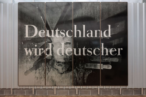 A multi-panel wall installation features a large-format black-and-white photograph with a heavily weathered, scratched surface. Across the image, the text “Deutschland wird deutscher” is written in large white letters. Behind the text, a vague figure can be made out.