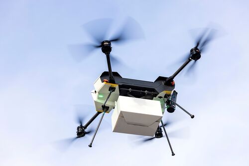 A delivery drone with four rotors is flying against a clear sky. A white parcel is attached beneath the central body. The rotors are spinning, and the drone has technical components and sensors on the front.
