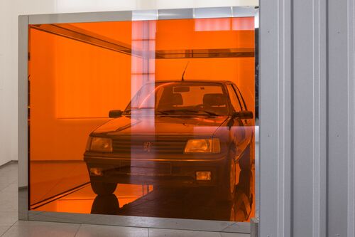 Ein Auto steht in einem Ausstellungsraum hinter einer orangefarbenen, transparenten Wand oder Folie. Durch die intensive orange Tönung erscheinen Fahrzeug und Innenraum nur schemenhaft.