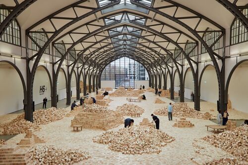Große begehbare Kunstinstallation aus tausenden hellen Holzblöcken in einer historischen Ausstellungshalle mit hohen Stahlbögen und Oberlichtern. Die Holzblöcke sind zu Wegen, Terrassen, Mauern und Hügeln angeordnet. Mehrere Besuchende bewegen sich durch die Installation, stapeln Holzblöcke um oder betrachten die verschiedenen Strukturen.