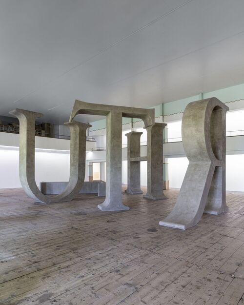 Großformatige Skulptur aus monumentalen, steinfarbenen Buchstaben H, R, T und U in einer weitläufigen Innenhalle. Die übergroßen Zeichen sind so angeordnet, dass man zwischen ihnen hindurchgehen kann. Im Hintergrund sind hohe Fenster. Eine einzelne Person geht in der Halle an den Buchstaben vorbei.