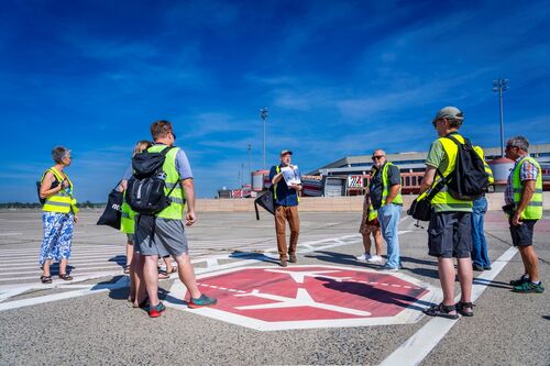 View across the airfield to the building of the former Tegel Airport. In the centre of the picture, participants in a guided tour are wearing yellow high-visibility waistcoats and looking towards the building.