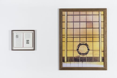 Zwei gerahmte Werke hängen an einer weißen Ausstellungswand. Rechts zeigt eine großformatige Fotografie oder Druckgrafik eine architektonische Glasfassade mit einem kreisförmigen Element vor einem gelblich eingefärbten Hintergrund. Links befindet sich ein kleineres gerahmtes Werk mit Text und Bild auf einer Buch- oder Dokumentenseite.