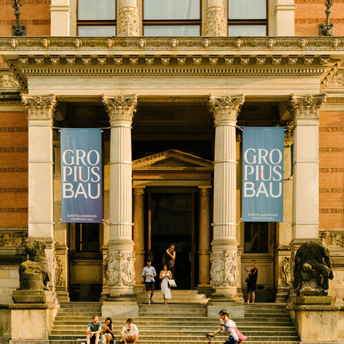 Der Eingang des Martin-Gropius-Baus mit seinen markanten Säulen und Aufgang. Zwei große blaue Banner mit dem Titel "Gropius Bau" hängen zwischen den Säulen.