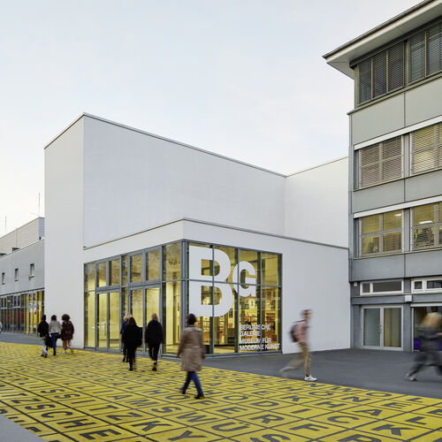 The entrance to the Berlinische Galerie. In front of it, a few people walk across the iconic yellow and black field of letters.