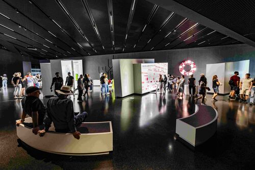 Ein großer, atmosphärisch beleuchteter Ausstellungsraum mit zahlreichen Menschen, die Schauwände ansehen, weiterlaufen oder im Vordergrund sitzen