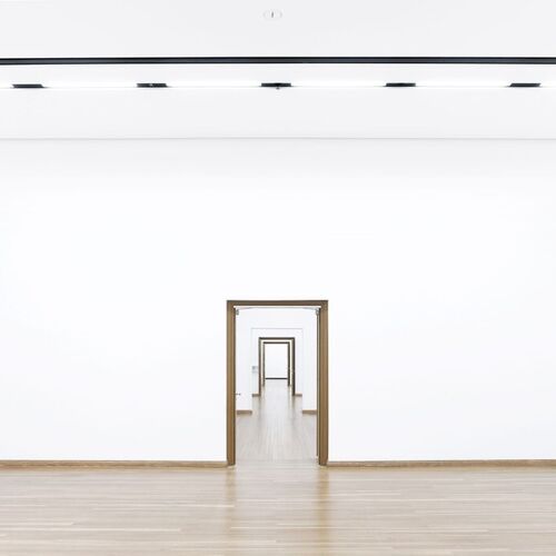 Innenansicht der leeren Ausstellungsräume des Gropius Baus: Ein langer, schmaler Flur mit weißen Wänden und einem hellen Holzboden. Mehrere Türen in der Ferne öffnen sich zu weiteren Räumen.