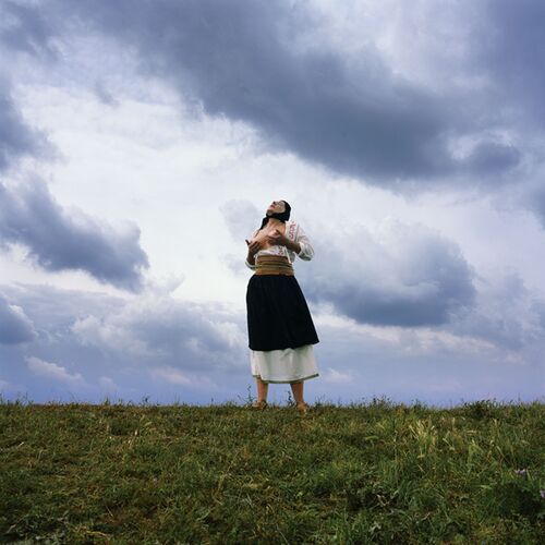 A single person stands in a meadow beneath a vast, dramatically overcast sky. The person is wearing a multi-layered, traditional-looking outfit consisting of a long, dark skirt and layers of light-coloured fabric. Their arms are bent and held in front of their torso.