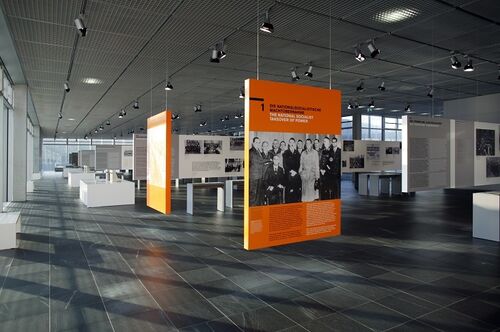 In der Ausstellungshalle der Topographie des Terrors stehen großformatige, orangefarbene Tafeln mit historischen Fotografien und erläuterndem Text zu den Verbrechen des nationalsozialistischen Regimes.