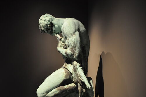 Eine knieende, bronzefarbende Statur in der Ausstellung. Ansicht von der Seite.