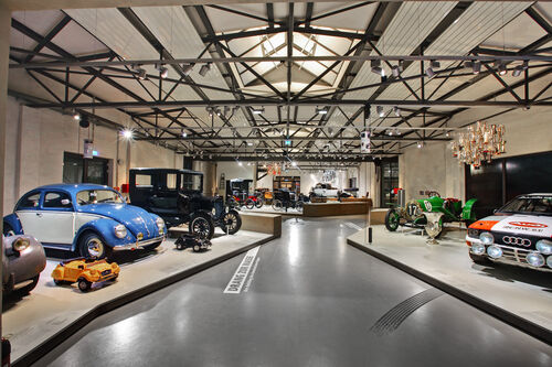 Blick in die Straßenverkehr-Ausstellung: Eine große Halle mit Stahlkonstruktion an der Decke. Ein breiter Gang zwischen niedrigen Podesten mit verschiedenen Fahrzeugen und Objekten.