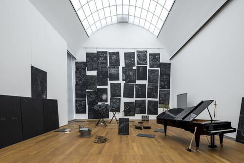 Ausstellungsansicht mit dem Werk „Das Kapital“ von Joseph Beuys. Schwarze mit Kreide beschriftete Tafeln an den Wänden, verschiedene Objekte auf dem Boden und rechts ein Konzertflügel, an dem eine Axt lehnt.