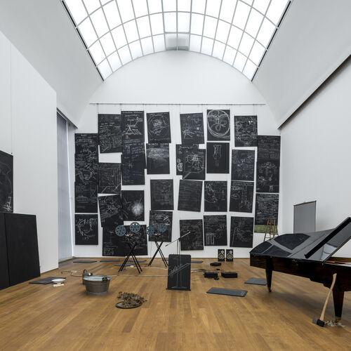 Ausstellungsansicht mit dem Werk „Das Kapital“ von Joseph Beuys. Schwarze mit Kreide beschriftete Tafeln an den Wänden, verschiedene Objekte auf dem Boden und rechts ein Konzertflügel, an dem eine Axt lehnt.