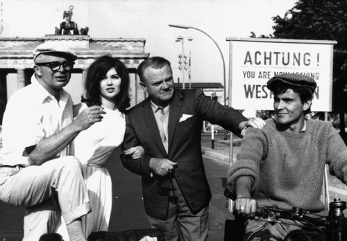 In einer historischen Aufnahme sind vier Personen vor dem Brandenburger Tor zu sehen. An der rechten Seite steht ein Schild, auf dem auf das Verlassen West-Berlins hingewiesen wird.