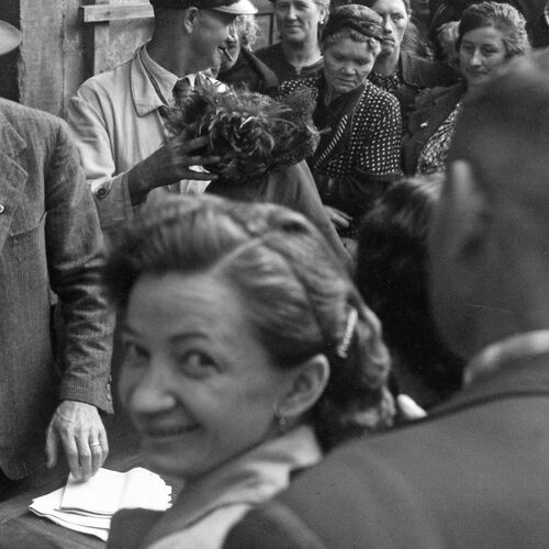 Historische Schwarz-Weiß-Fotografie einer Menschenmenge im öffentlichen Raum. Im Vordergrund blickt eine Person über die Schulter in Richtung Kamera, während zahlreiche weitere Personen dicht gedrängt um einen Tisch oder eine Ausgabestelle versammelt sind. Links steht eine Person mit Hut und Anzug neben einem Stapel Papier.