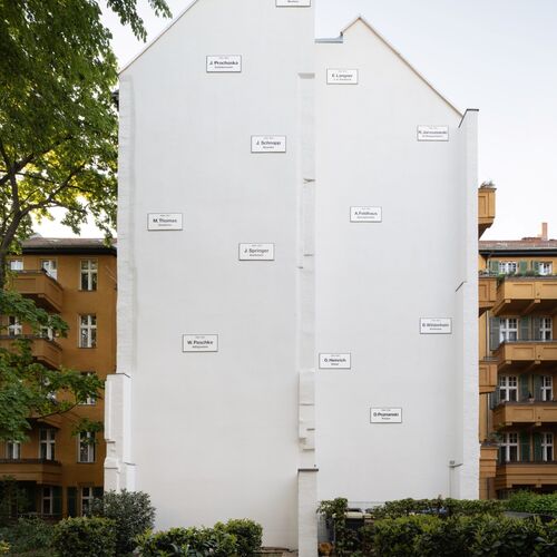 Foto der Installation "Missing House " von Christian Boltanski. Mittig eine weiße Brandmauer an der 11 weiße kleinere Schilder mit schwarzer Schrift angebracht sind.