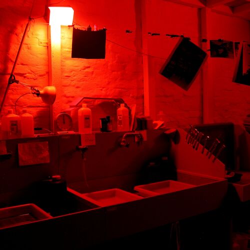 Ein dunkler Raum, der in tiefrotes Licht getaucht ist und wie eine analoge Fotolabor-Dunkelkammer wirkt. Im Vordergrund stehen mehrere rechteckige Entwicklerwannen in einer langen Spüle. Dahinter befinden sich verschiedene Flaschen und Behälter mit Fotochemikalien auf einer Ablage. An der Wand hängen Fotopapiere und Clips an gespannten Leinen.&nbsp;