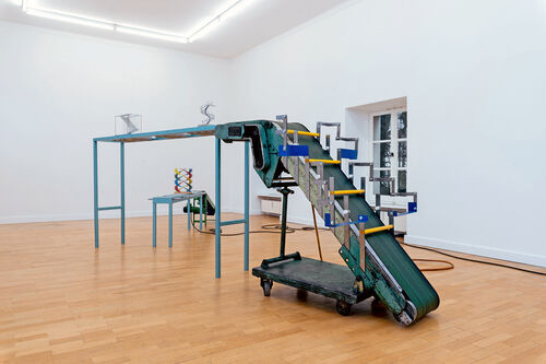 Kunstinstallation in einem hellen Raum. Im Zentrum steht eine große, industrielle Förderbandanlage aus Metall. Das Förderband ist in einem Winkel nach oben geführt und geht dann waagerecht weiter. 