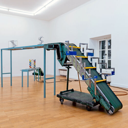 Kunstinstallation in einem hellen Raum. Im Zentrum steht eine große, industrielle Förderbandanlage aus Metall. Das Förderband ist in einem Winkel nach oben geführt und geht dann waagerecht weiter. 