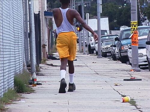 Ein Filmstill aus "Niagara" (2005) von Mark Bradford. Von hinten: Eine Person in einer kurzen, gelben Hose läuft durch eine Straße. Autos parken neben dem etwas vermüllten Bürgersteig.