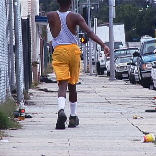 Ein Filmstill aus "Niagara" (2005) von Mark Bradford. Von hinten: Eine Person in einer kurzen, gelben Hose läuft durch eine Straße. Autos parken neben dem etwas vermüllten Bürgersteig.
