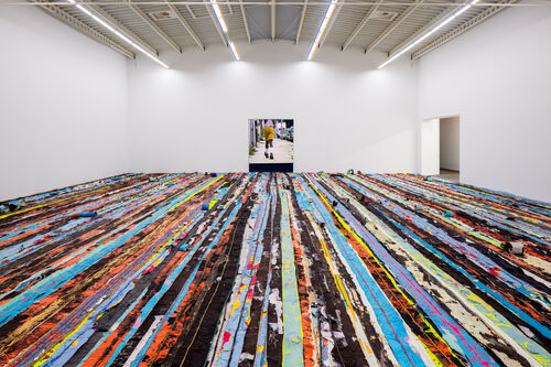 Viele bunte Farbstreifen, die den Boden einer großen Halle bedecken und eine Videoleinwand mit einer männlichen Person in der Ausstellungsansicht Mark Bradford Keep Walking