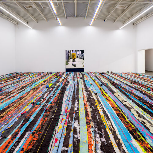 Viele bunte Farbstreifen, die den Boden einer großen Halle bedecken und eine Videoleinwand mit einer männlichen Person in der Ausstellungsansicht Mark Bradford Keep Walking
