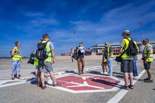 Blick über das Flugfeld zum Gebäude des ehemaligen Flughafens Tegel. In der Bildmitte stehen Teilnehmer*innen einer Führung in gelben Warnwesten und schauen gen Gebäude.