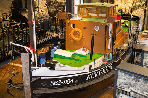 Der schwarz, grün, braune Schlepp- und Schubdampfer "Kurt Heinz" steht in der Schifffahrtsausstellung des Deutschen Technikmuseum. Kinder sind auf dem Deck.