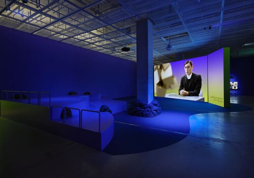 Eine große Videoleinwand mit einer männlichen Person befindet sich in einem dunkelblau schimmernden Raum mit Sitzgelegenheiten in der Ausstellungsansicht Ayoung Kim