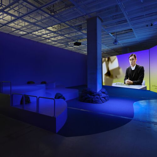 Eine große Videoleinwand mit einer männlichen Person befindet sich in einem dunkelblau schimmernden Raum mit Sitzgelegenheiten in der Ausstellungsansicht Ayoung Kim