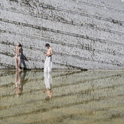 zwei einander zugewandt stehende und transparent gekleidete Männer vor einer bildfüllenden Mauerstruktur spiegeln sich in einer Wasserfläche während einer Performance von Klara Hosnedlova
