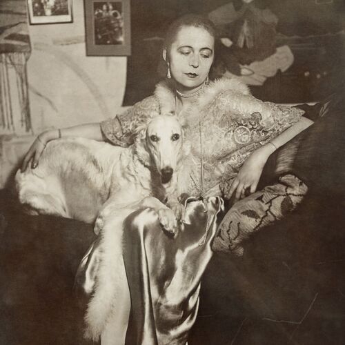 Selbstportrait der Künstlerin Marta-Astfalck-Vietz als Schwarz-Weiß-Fotografie. Sie sitzt in einem glänzenden Kleid mit Pelzbesatz auf einem Sofa. Auf ihrem Schoß sitzt ein Hund mit hellem Fell. Im Hintergrund sind Gemälde und dekorative Elemente zu sehen.