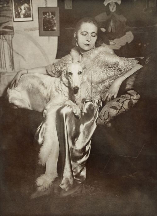 Selbstportrait der Künstlerin Marta-Astfalck-Vietz als Schwarz-Weiß-Fotografie. Sie sitzt in einem glänzenden Kleid mit Pelzbesatz auf einem Sofa. Auf ihrem Schoß sitzt ein Hund mit hellem Fell. Im Hintergrund sind Gemälde und dekorative Elemente zu sehen.