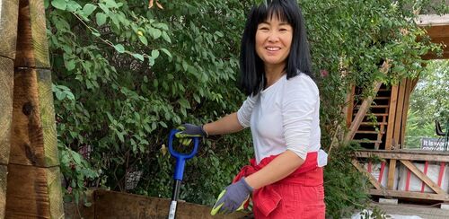 Ein Foto der Künstlerin Ayumi Matsuzaka mit Gartenhandschuhen vor einer grünen Hecke. Sie hält einen Stiel eines Arbeitsgeräts in der Hand und blickt uns lächelnd an.