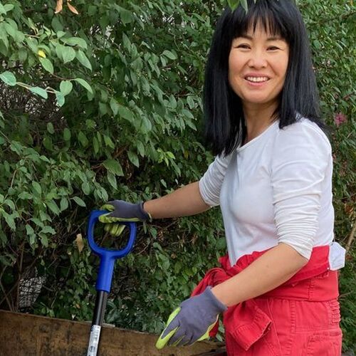 Ein Foto der Künstlerin Ayumi Matsuzaka mit Gartenhandschuhen vor einer grünen Hecke. Sie hält einen Stiel eines Arbeitsgeräts in der Hand und blickt uns lächelnd an.