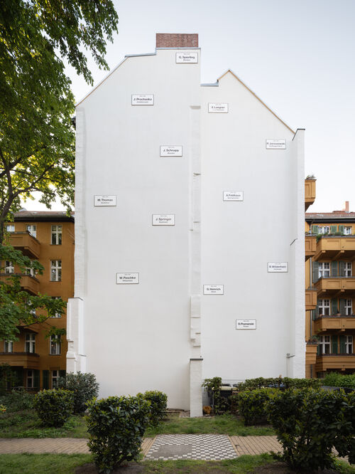 Foto der Installation "Missing House " von Christian Boltanski. Mittig eine weiße Brandmauer an der 11 weiße kleinere Schilder mit schwarzer Schrift angebracht sind.