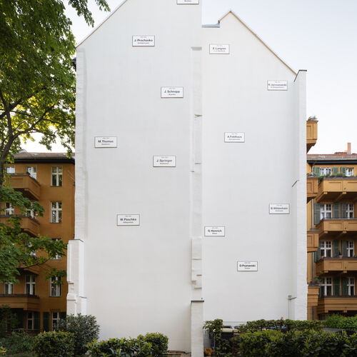 Foto der Installation "Missing House " von Christian Boltanski. Mittig eine weiße Brandmauer an der 11 weiße kleinere Schilder mit schwarzer Schrift angebracht sind.