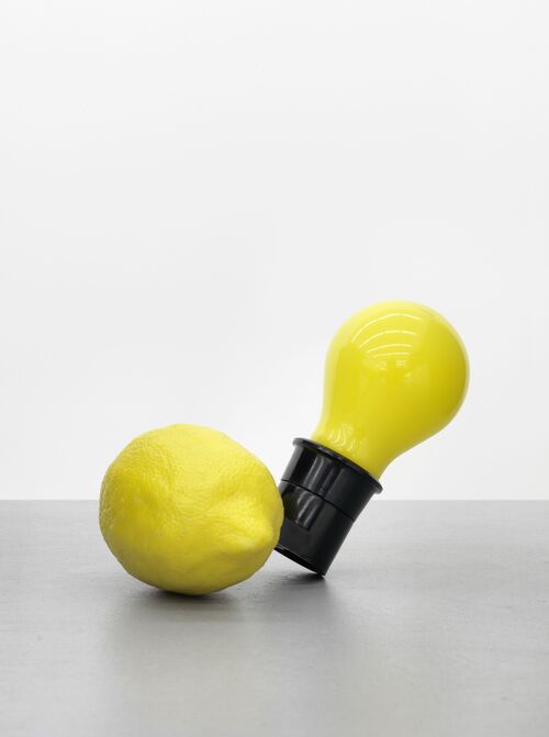 Das Werk Capri-Batterie von Joseph Beuys. Eine gelbe Glühbirne verbunden mit einer liegenden gelben Zitrone vor einem neutralen hellgrauen Hintergrund.