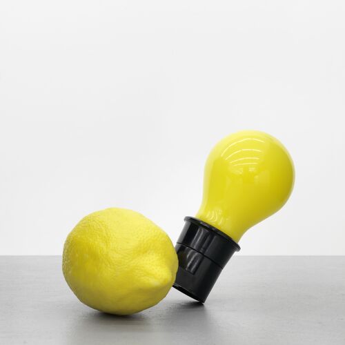 Das Werk Capri-Batterie von Joseph Beuys. Eine gelbe Glühbirne verbunden mit einer liegenden gelben Zitrone vor einem neutralen hellgrauen Hintergrund.